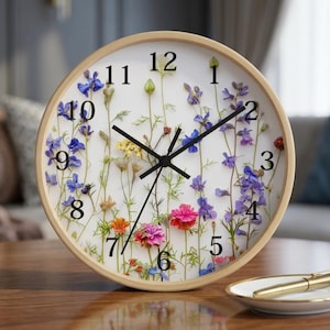 Op de afbeelding: Een ronde klok met een licht houten frame. De witte wijzerplaat heeft zwarte cijfers en wijzers. Kleurrijke bloemen in blauw, paars, roze en geel zijn op de wijzerplaat geplaatst. De klok staat op een houten oppervlak.