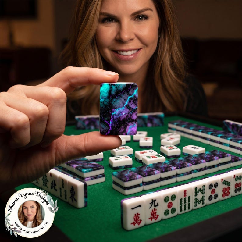 Puede incluir: Primer plano de una ficha de Mahjong con colores turquesa, morado y negro, sostenida por una persona. Otras fichas de Mahjong est&aacute;n sobre una superficie verde. El juego incluye fichas blancas con marcas rojas y verdes.