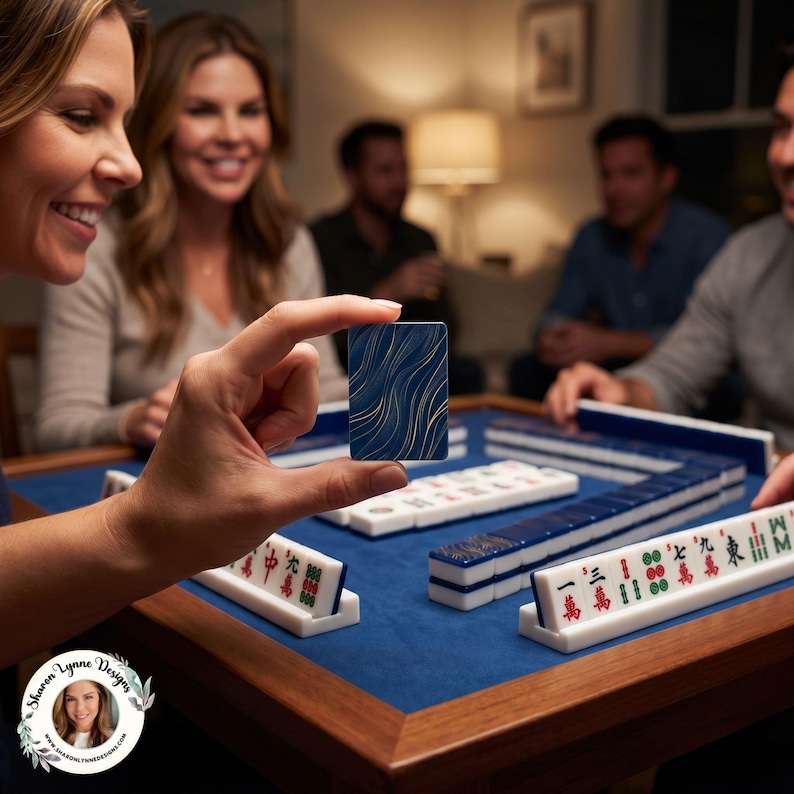 May include: A rectangular mahjong tile held between fingers, featuring a dark blue background with gold wavy lines. The game set includes white tiles with red, green, and black Chinese characters arranged on a blue felt surface on a wooden table.