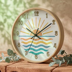 May include: A round wall clock with a light wood frame. The clock face features a sunrise design with yellow, orange, and blue waves. Black hands indicate the time. The clock is approximately 25 cm in diameter.