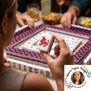 K&ouml;nnte beinhalten: Eine Person h&auml;lt einen wei&szlig;en Mahjong-Stein mit einem roten Drachenmuster. Der Stein ist Teil eines Mahjong-Sets mit rosa und schwarz gemusterten R&auml;ndern. Weitere Steine und Spielelemente sind auf dem Tisch sichtbar.
