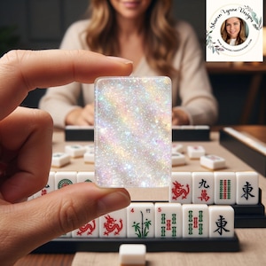 May include: A rectangular mahjong tile featuring a clear, iridescent, and glittery finish that reflects light with rainbow hues. The tile is held upright, showcasing a dense, sparkling texture throughout its surface, distinct from the standard white tiles.