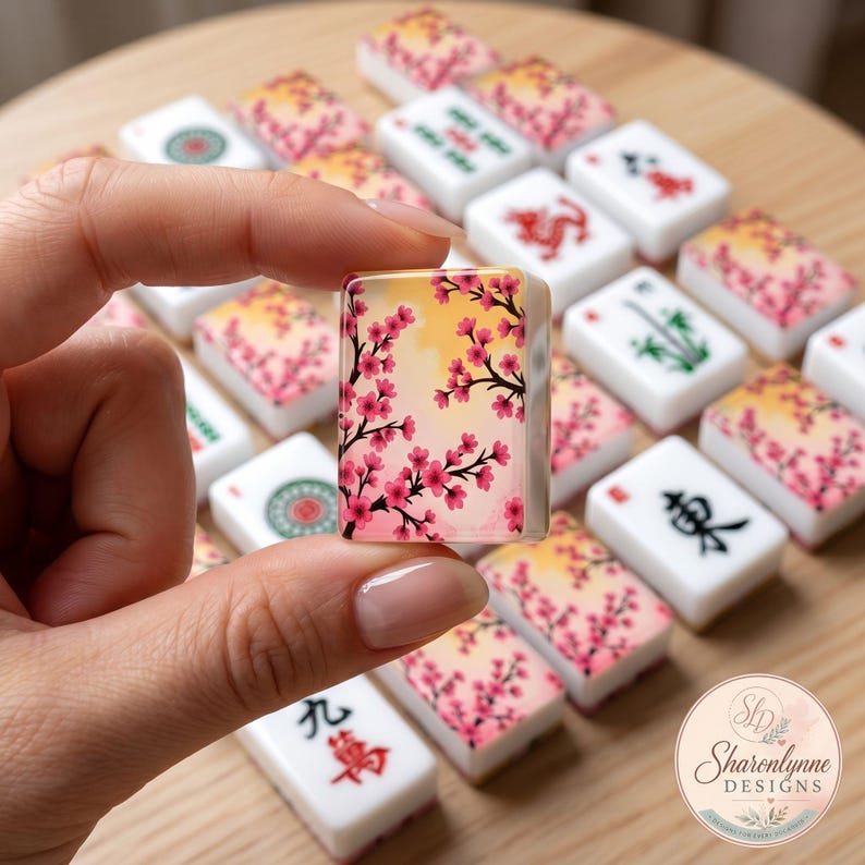 May include: A rectangular mahjong tile featuring a printed design of pink cherry blossom branches against a soft yellow and white background, held up among a collection of various other mahjong tiles with traditional symbols and floral patterns.