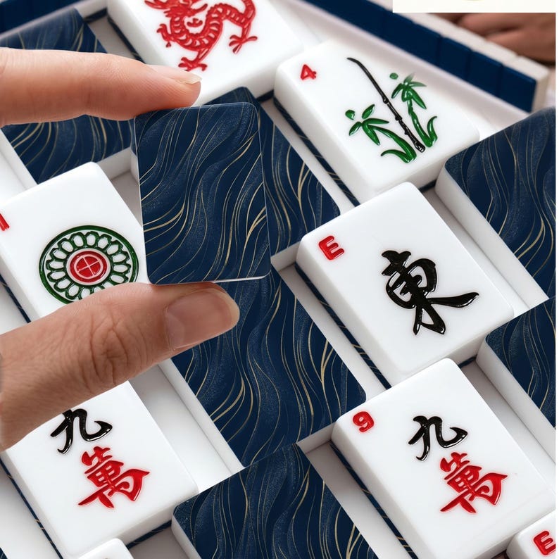 May include: A set of Mahjong tiles featuring white faces with red, green, and black traditional characters and symbols, alongside dark blue tiles with a gold wavy line pattern. One dark blue tile is held up to display the back design clearly.