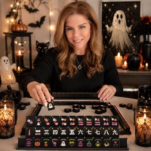 A smiling woman sits at a table arranged with the black mahjong tile set and Halloween decorations.