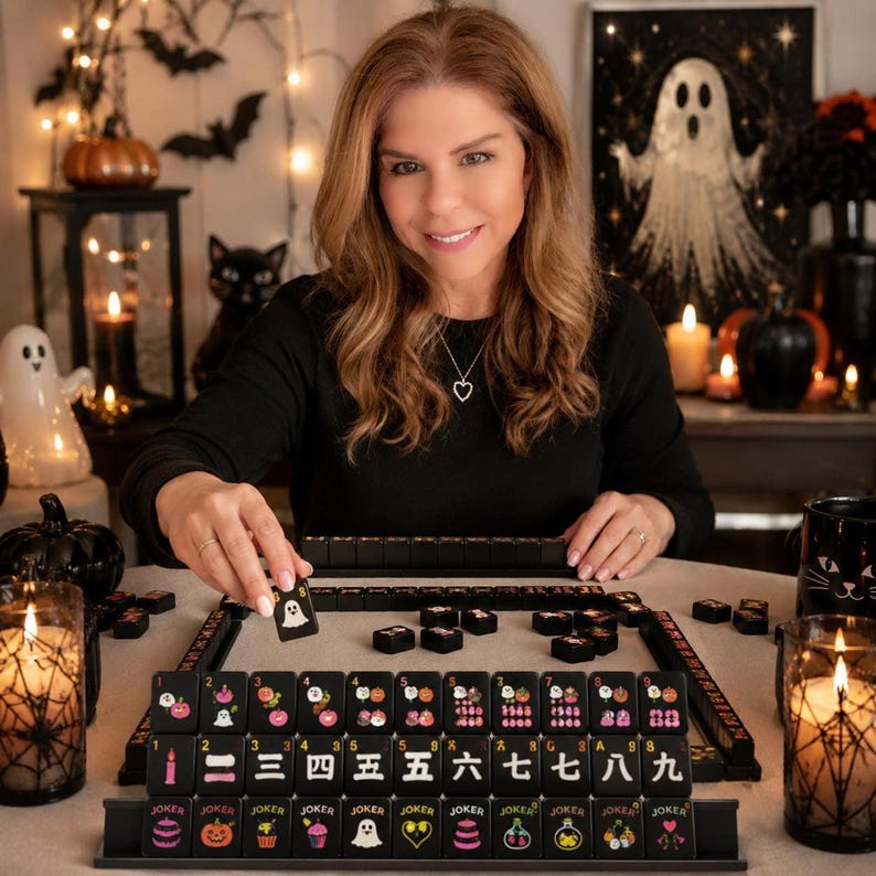 A smiling woman sits at a table arranged with the black mahjong tile set and Halloween decorations.