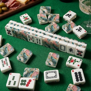 May include: A collection of white and marble-patterned Mahjong tiles scattered on a green surface. The tiles feature various symbols, numbers, and Chinese characters in black and red. Some tiles have floral designs.