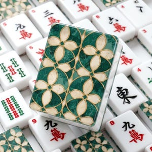 May include: A rectangular mahjong tile featuring a decorative back with a green marble-like pattern and cream-colored floral shapes outlined in gold. The tile is surrounded by other white mahjong tiles displaying various red and black Chinese characters.