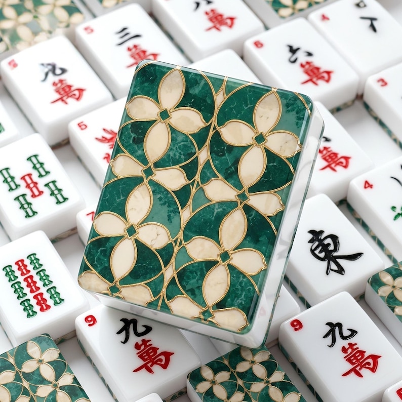May include: A rectangular mahjong tile featuring a decorative back with a green marble-like pattern and cream-colored floral shapes outlined in gold. The tile is surrounded by other white mahjong tiles displaying various red and black Chinese characters.