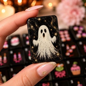 A hand holds a single black mahjong tile featuring a white ghost illustration surrounded by gold sparkle effects.