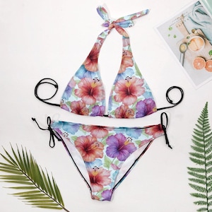 A floral halter bikini set with tie-side bottoms is laid flat on a white surface, with a green fern leaf and a magazine in the background.