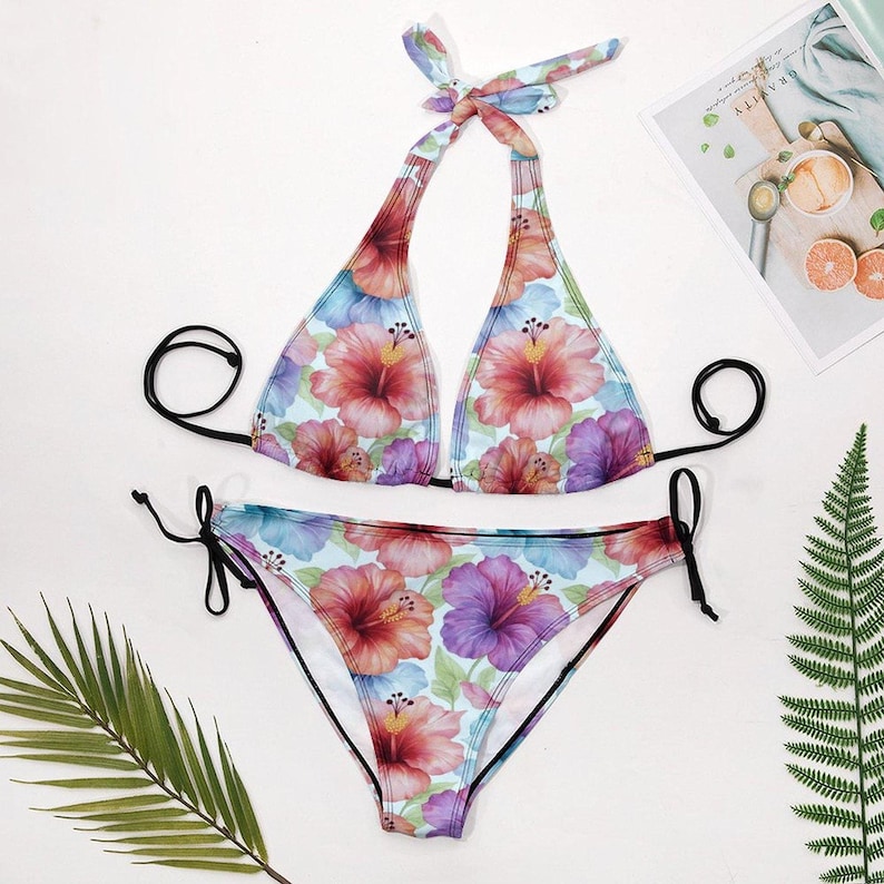 A floral halter bikini set with tie-side bottoms is laid flat on a white surface, with a green fern leaf and a magazine in the background.