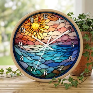 May include: A round wall clock with a light wood frame featuring a stained glass style design of a sunset over the ocean. The clock face displays a yellow sun, pink clouds, and blue water with black numerals from 1 to 12 and white clock hands.
