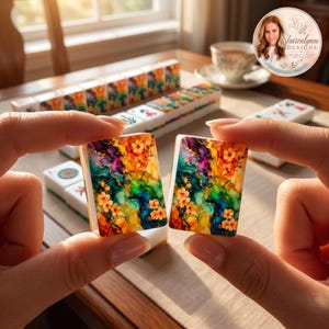 May include: Two rectangular mahjong tiles featuring a vibrant alcohol ink design with swirling patterns of blue, green, purple, and yellow, accented by small orange flowers. The tiles have a white base and are held up against a blurred background.