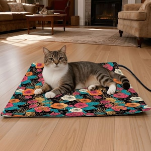 May include: A tabby cat rests on a rectangular pet heating pad. The pad features a black background with a colorful floral pattern of roses in shades of orange, pink, teal, and white. A black cord extends from the pad.