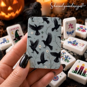 May include: A rectangular tile featuring a light blue background with a repeating pattern of black crows in flight, delicate thin branches, and small white flowers. The tile has smooth, rounded edges and a glossy finish, held in a hand.