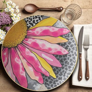 May include: A decorative plate with a large, stylized pink and yellow flower design. The plate has a gold rim and a gray background with a leopard print pattern. A wooden spoon, glass, and silverware are also visible.