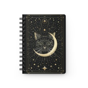 Front cover of a black spiral notebook featuring a gold celestial cat illustration with a crescent moon and stars.