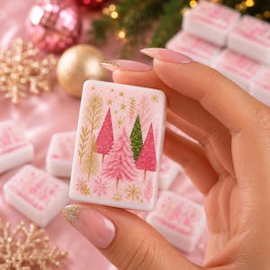 May include: A rectangular mahjong tile featuring a light pink background with a festive design of pink, green, and gold Christmas trees, gold stars, and gold snowflakes. The tile has a smooth, glossy finish and is held between two fingers.