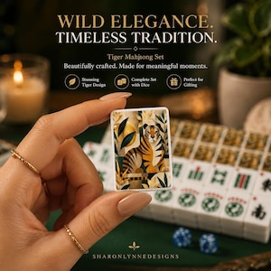 May include: A rectangular white mahjong tile featuring a graphic illustration of a tiger surrounded by stylized foliage and a golden sun. The tile is held in front of a complete set of matching mahjong tiles and two blue dice on a green surface.