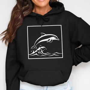 May include: Black hooded sweatshirt featuring a white graphic of a dolphin leaping over waves within a white square. The sweatshirt has a front pocket and a drawstring hood. The design is simple and clean.