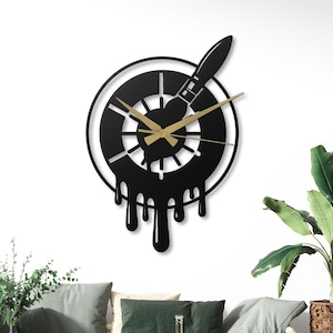 May include: Black metal wall clock featuring a paintbrush design. The clock has gold-colored hands and a unique dripping paint effect. The clock is mounted on a white wall, adding a modern touch to any room.