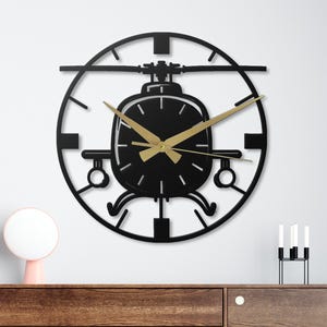 May include: Black metal helicopter wall clock with gold-colored hands. The clock features a circular design with a helicopter silhouette in the center. The clock is mounted on a white wall above a wooden cabinet.