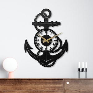 Op de afbeelding: Zwarte metalen anker wandklok met een ronde wijzerplaat en goudkleurige wijzers. De klok is ontworpen met een nautisch thema, met een touwdetail en Romeinse cijfers. De klok is gemonteerd op een witte muur.