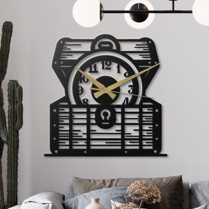 Könnte beinhalten: Eine schwarze Metall-Wanduhr in Form einer offenen Schatzkiste mit einem Zifferblatt im Inneren. Die Uhr hat goldfarbene Zeiger und ein weißes Zifferblatt mit schwarzen Ziffern. Die Uhr ist an einer weißen Wand befestigt.