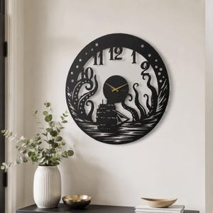 Op de afbeelding: Een ronde zwarte wandklok met een uitgesneden ontwerp van een zeilschip dat wordt aangevallen door een grote octopus. De wijzerplaat heeft zwarte cijfers van één tot twaalf en dunne goudkleurige wijzers op een zwarte ronde achtergrond.