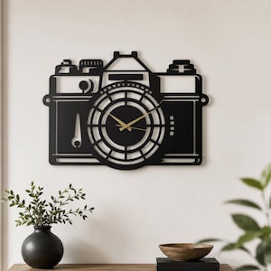 Pode incluir: Um relógio de parede preto em forma de câmara vintage com um design central de lente circular. O relógio apresenta ponteiros finos de cor dourada para as horas, minutos e segundos no centro. O corpo da câmara é uma silhueta de metal plana.