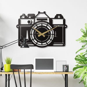 May include: Black metal wall clock in the shape of a vintage camera. The clock face is circular with gold-colored hands. The clock is mounted on a white wall above a wooden desk with a black chair.