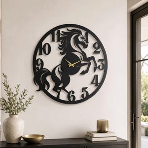 May include: A circular black wall clock featuring a cutout silhouette of a rearing horse in the center. The clock face includes large black numerals from one to twelve integrated into the circular frame, paired with thin gold-colored clock hands.
