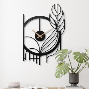 May include: Black metal wall clock with a circular design featuring a stylized leaf and geometric shapes. The clock has gold-colored hands and a black face.