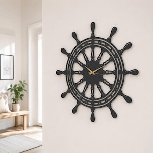 May include: A black wall clock shaped like a ship's steering wheel with twelve spokes radiating from a central hub. The clock features thin, gold-colored hour and minute hands positioned in the center of the circular frame, mounted on a wall.