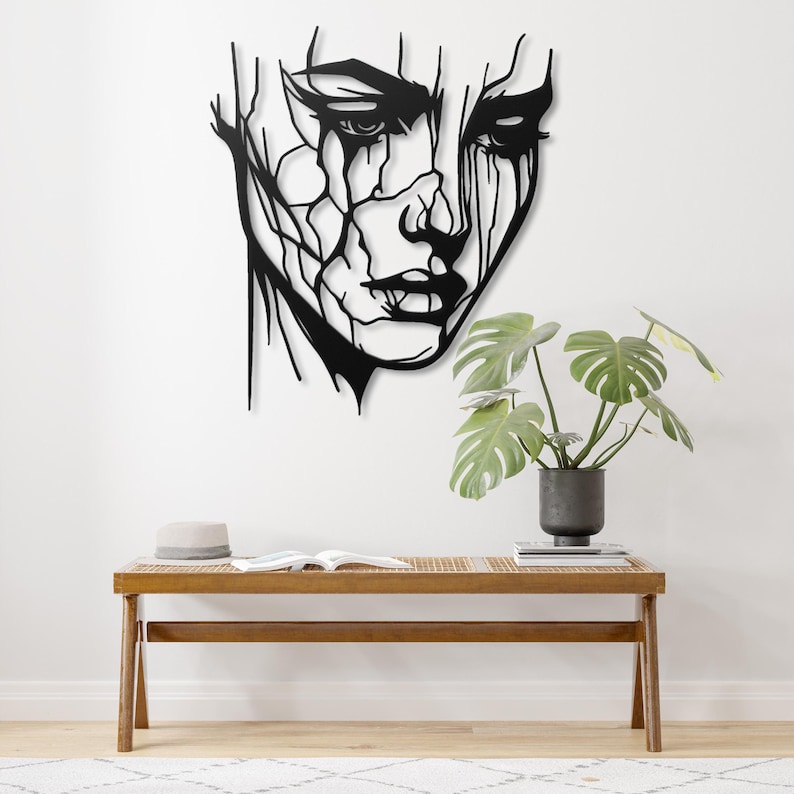 K&ouml;nnte beinhalten: Schwarze Wandkunst aus Metall mit einem stilisierten abstrakten Gesicht mit einem verwitterten Look. Das Kunstwerk ist an einer wei&szlig;en Wand &uuml;ber einer Holzbank angebracht.