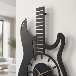 K&ouml;nnte beinhalten: Eine mattschwarze Wanduhr in Form einer E-Gitarren-Silhouette. Die Uhr verf&uuml;gt &uuml;ber einen langen Hals mit horizontalen Ausschnitten, einen kreisf&ouml;rmigen K&ouml;rper mit strahlenf&ouml;rmigen Stundenmarkierungen und d&uuml;nne goldene Zeiger in der Mitte.