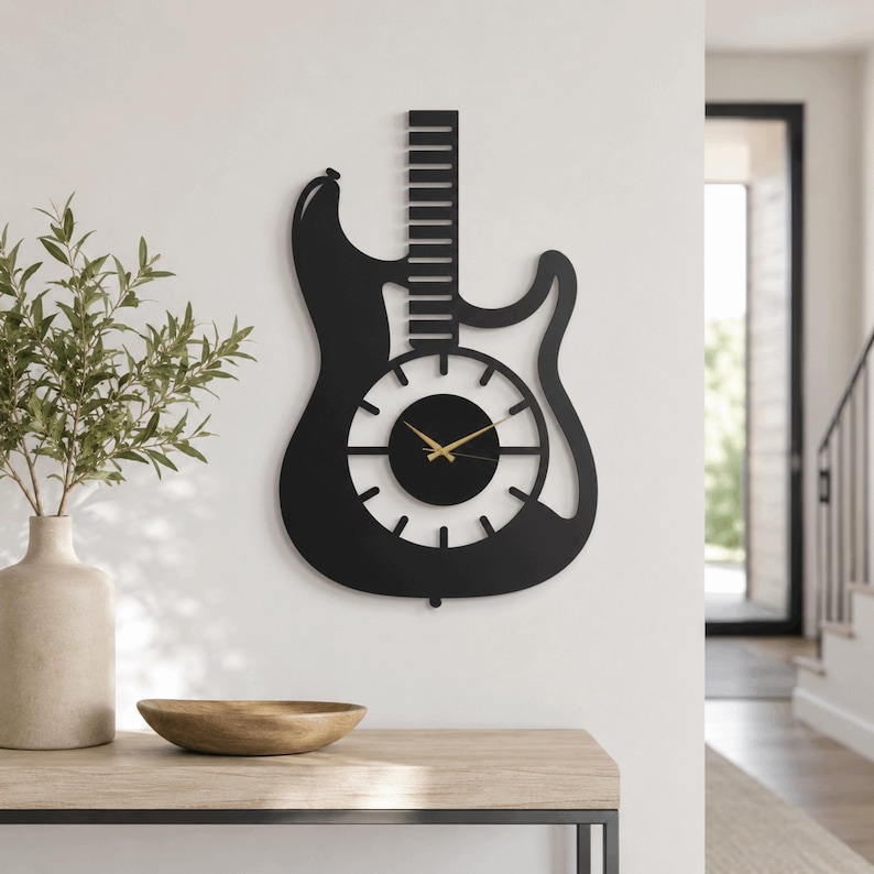K&ouml;nnte beinhalten: Eine schwarze Wanduhr in Form einer E-Gitarren-Silhouette. Die Uhr verf&uuml;gt &uuml;ber ein zentrales, kreisf&ouml;rmiges Zifferblatt mit d&uuml;nnen, strahlenf&ouml;rmigen Stundenmarkierungen und zwei goldfarbenen Zeigern. Der Gitarrenhals ragt als vertikaler Balken nach oben.