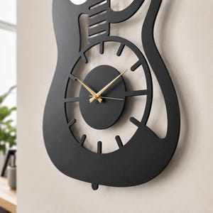 K&ouml;nnte beinhalten: Eine mattschwarze Wanduhr in Form eines E-Gitarrenkorpus. Die Uhr verf&uuml;gt &uuml;ber ein kreisf&ouml;rmiges Zentrum mit d&uuml;nnen, goldfarbenen Zeigern f&uuml;r Stunden, Minuten und Sekunden. Der &auml;u&szlig;ere Rahmen ist eine massive schwarze Silhouette einer Gitarre.