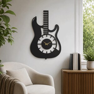K&ouml;nnte beinhalten: Eine mattschwarze Wanduhr in Form einer E-Gitarren-Silhouette. Die Uhr verf&uuml;gt &uuml;ber ein zentrales, kreisf&ouml;rmiges Zifferblatt mit zw&ouml;lf strahlenf&ouml;rmigen Stundenmarkierungen und zwei d&uuml;nnen, goldfarbenen Zeigern. Der Gitarrenhals ragt vertikal empor.