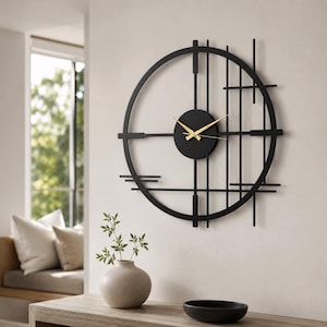 Könnte beinhalten: Eine große, kreisförmige Wanduhr mit einem schwarzen Metallrahmen und geometrischen vertikalen sowie horizontalen Linien. In der Mitte befindet sich ein massives, schwarzes Zifferblatt mit dünnen, goldfarbenen Stunden- und Minutenzeigern.