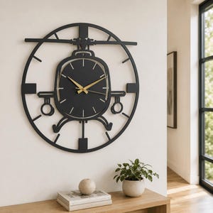 Op de afbeelding: Een ronde wandklok met een zwart metalen frame in de vorm van een helikoptersilhouet. De centrale wijzerplaat is zwart met dunne goudkleurige uur- en minuutwijzers. Het ontwerp bevat gestileerde rotorbladen en landingsgestellen.