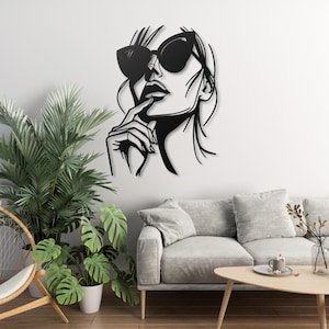 Puede incluir: Arte de pared de metal negro con una silueta estilizada de una mujer con gafas de sol mirando hacia arriba. El cabello de la mujer es largo y fluido, y su mano descansa sobre su barbilla.