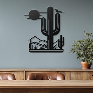 May include: Black metal wall art depicting a desert landscape with a large cactus, smaller cacti, mountains, and a bird perched on the cactus. The art is mounted on a light grey wall.
