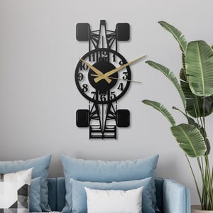 May include: Black metal wall clock shaped like a Formula 1 race car. The clock face is round with gold hands and black numerals. The clock is mounted on a light gray wall.