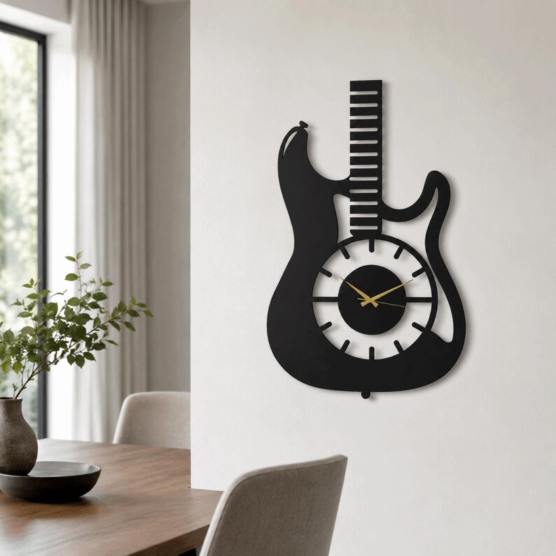 K&ouml;nnte beinhalten: Eine mattschwarze Wanduhr in Form einer E-Gitarre. Die Uhr verf&uuml;gt &uuml;ber einen langen Hals mit horizontalen Bundlinien und ein kreisf&ouml;rmiges Zifferblatt, das in den Korpus integriert ist. Sie hat d&uuml;nne, goldfarbene Zeiger.