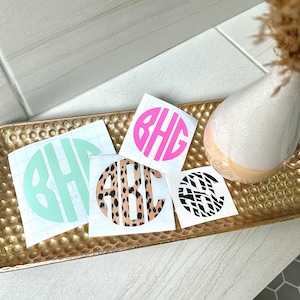May include: A collection of monogram decals in various colors and patterns. The decals feature initials in a circular design. Colors include mint green, hot pink, leopard print, and black and white geometric patterns. The decals are displayed on a gold tray.