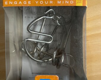 May include: A boxed metal puzzle game called "Puzzlers" with the text "Engage Your Mind." The puzzle is silver-colored and labeled "Fly Catcher." The box is orange and blue, with the word "Intermediate" on the top.