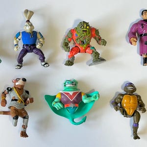 May include: A collection of nine action figures, including a green turtle, a purple-clad figure, a green figure with a cape, a brown figure with a sword, a green figure with a shell, a green figure with a shell, a green figure with a shell, a green figure with a shell, and a green figure with a shell.