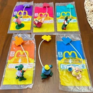 May include: A set of six McDonald's Dino-Motion Dinosaurs toys. Each toy is packaged in a clear plastic bag with a yellow and red cardboard backing. The toys feature different dinosaurs, including Charlene Sinclair, Baby Sinclair, and others. The toys are connected to a string that allows them to move when pulled.